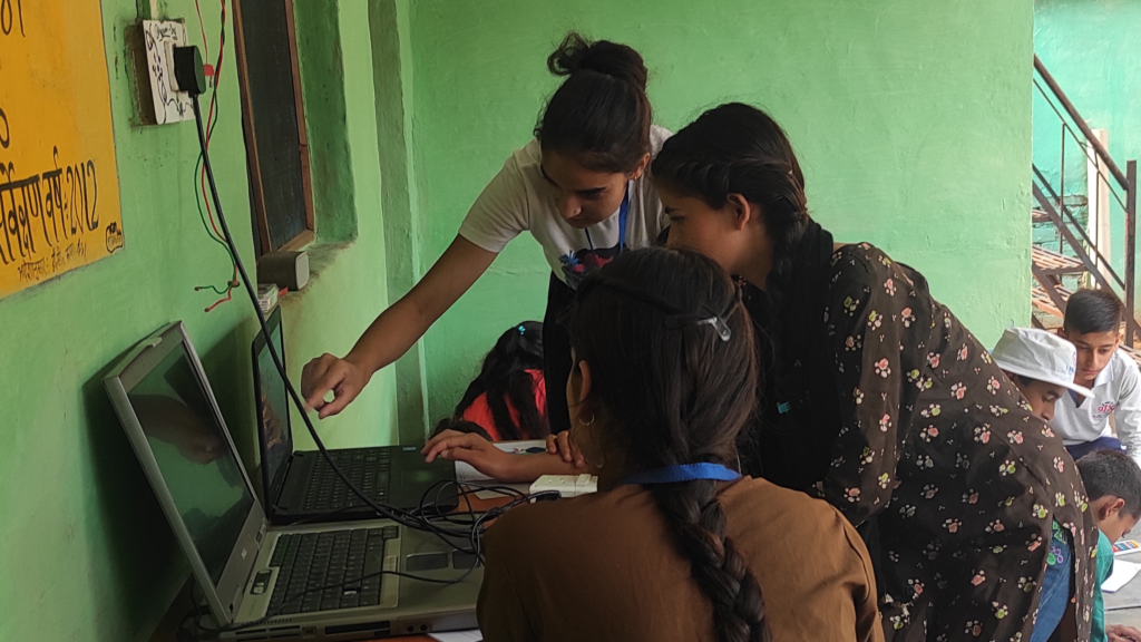 Female community leader guiding girls through a coding activity at the Rakkar village learning hub, with other children engaged in activities nearby.